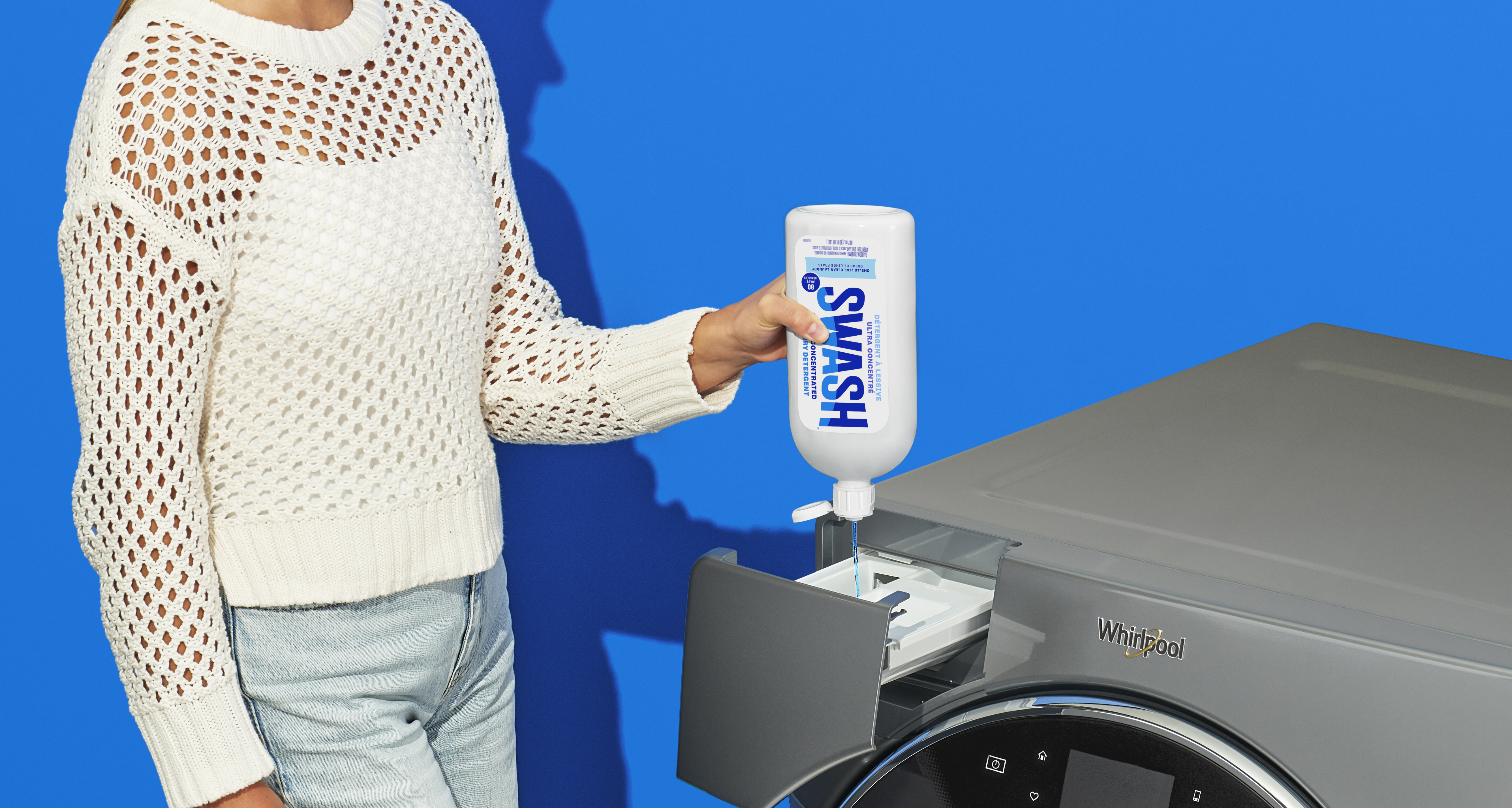 A woman squeezes detergent out of a bottle of Swash into the detergent dispenser of a Whirlpool Washing Machine in front of a blue background.
